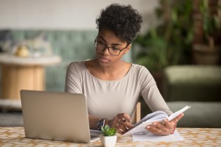 Young African American woman with glasses reading article on computer and taking notes