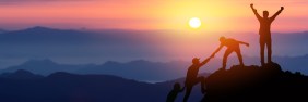 Group of friends climb mountain at sunrise while one friend celebrates at the top.