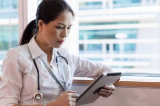 Asian-American female healthcare provider wearing a stethoscope and sitting in the window of a hospital reviewing her patients' charts on a tablet.