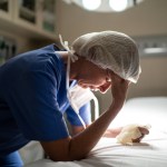 Stressed female nurse in PPE leans on bed and rubs forehead