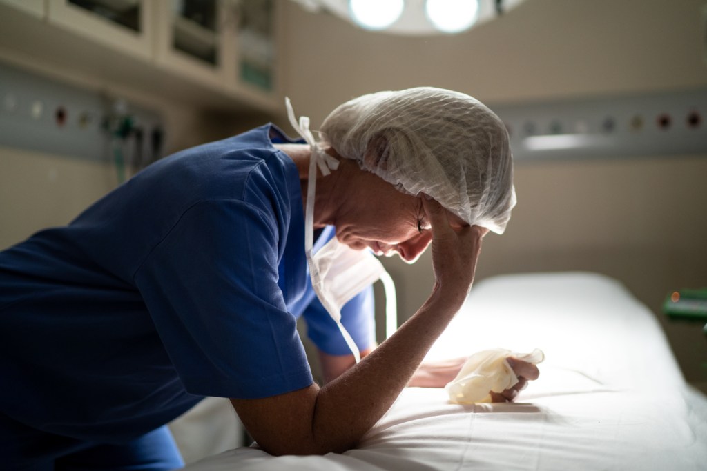 Stressed female nurse in PPE leans on bed and rubs forehead