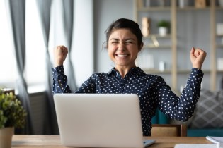 Young woman of color raising her arms in excitement while sitting in front of laptop.