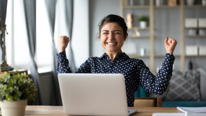 Young woman of color raising her arms in excitement while sitting in front of laptop.