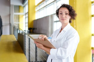 Young white female nurse with short brown curly hair wearing a white lab coat and holding a clipboard while standing in an open room with bright yellow walls and large windows.