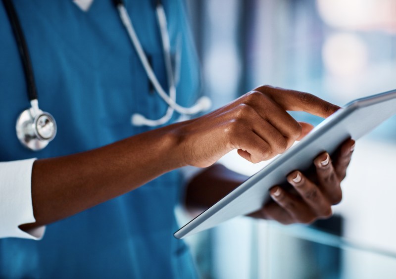 Lower half of African-American man wearing blue scrubs and a stethoscope scrolling through a document on a tablet.