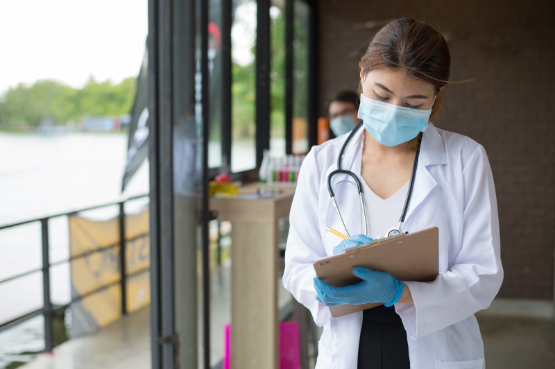 Young Asian-American female nurse wearing mask, white lab coat, and stethoscope taking notes on a clipboard in the middle of a hallway.