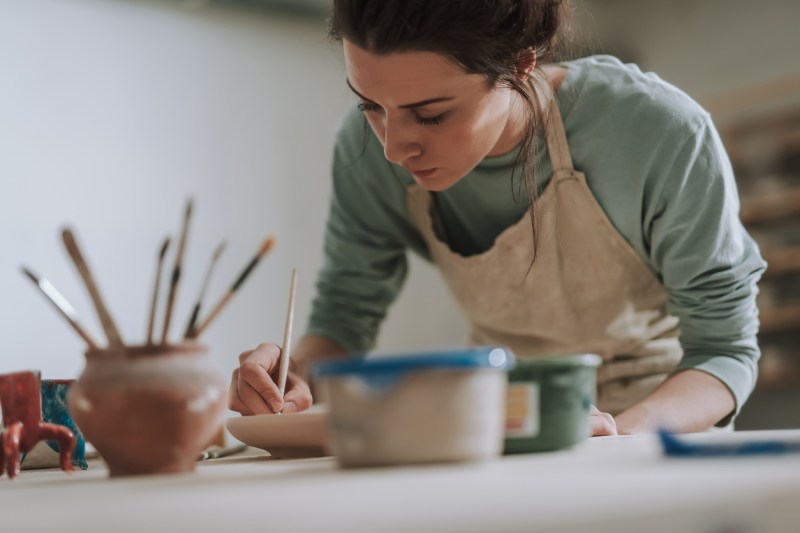 Young white woman in apron painting pottery in her workshop.