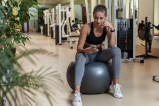 Young female physical therapist in activewear sits on gray yoga ball in the middle of a gym while checking her smartphone.