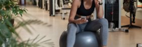 Young female physical therapist in activewear sits on gray yoga ball in the middle of a gym while checking her smartphone.
